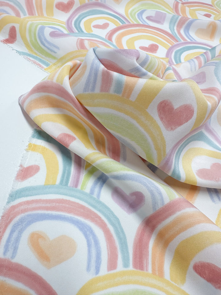A close-up of Super Cheap Fabrics' Printed Delsutered Satin - Unity - 155cm displays its lightweight, enchanting design with overlapping rainbows and hearts. Pastel yellow, pink, orange, and blue shades flow gracefully across the softly draped fabric.