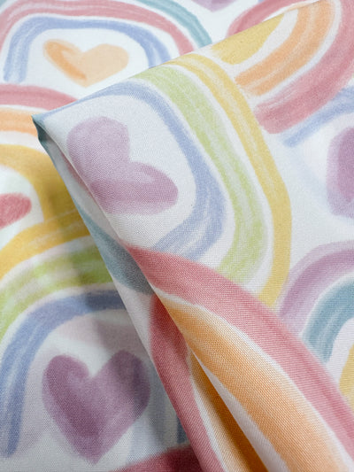 This lightweight fabric, the "Printed Delustered Satin - Unity - 155cm" by Super Cheap Fabrics, showcases pastel rainbow-colored hearts and arches in soft hues of pink, purple, yellow, green, and blue for a whimsical and playful look.