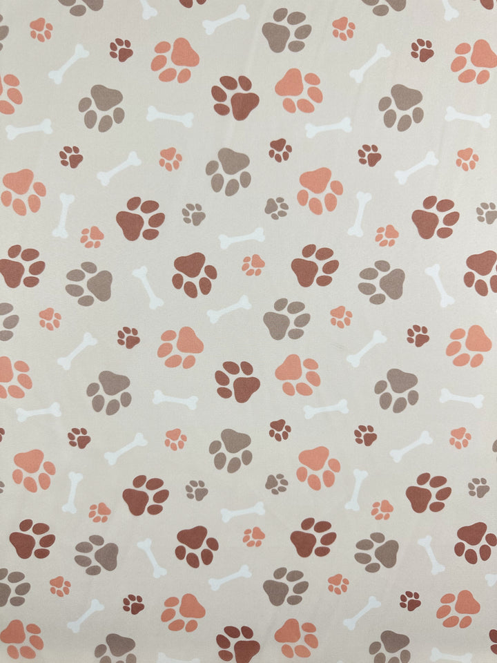 The Printed Delustered Satin - Pawsies by Super Cheap Fabrics features paw prints in brown, red, and peach with white dog bone shapes on a light beige background. Width: 155cm.