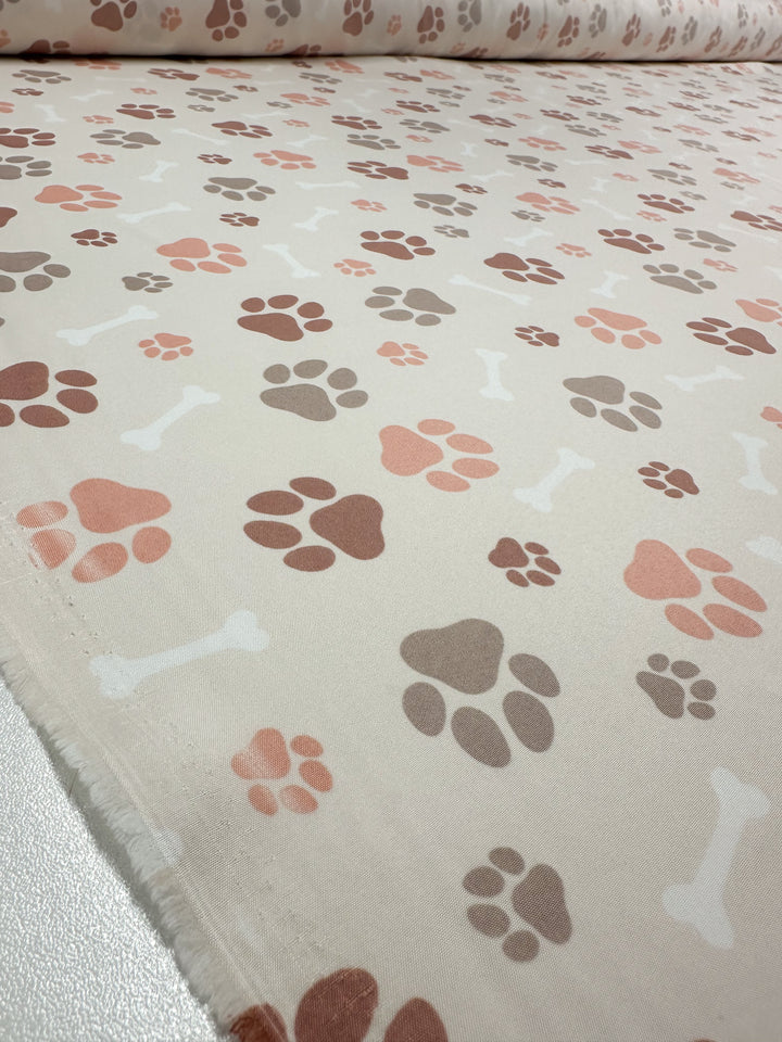 The "Printed Delustered Satin - Pawsies - 155cm" by Super Cheap Fabrics showcases a whimsical pattern of multicolored paw prints and bones on a light beige background.
