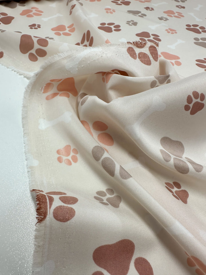 The Printed Delustered Satin - Pawsies by Super Cheap Fabrics, features charming pink and brown paw prints and bones on a soft, lightweight fabric. This 155cm wide satin is perfect for crafting elegant pieces or sewing projects full of charm.