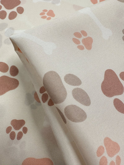 Close-up of Super Cheap Fabrics' Printed Delustered Satin - Pawsies, showcasing a whimsical pattern of brown, tan, and peach paw prints and dog bones on light beige. Slightly folded, it adds charm to these elegant 155cm pieces.