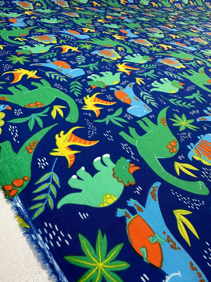 The Printed Delsutered Satin - Dino Jungle from Super Cheap Fabrics features playful green, orange, and blue dinosaurs among tropical leaves and white dots on blue fabric, blending whimsy with elegance.