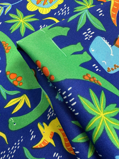 The Printed Delustered Satin - Dino Jungle from Super Cheap Fabrics features playful green, blue, and yellow dinosaurs mixed with green and yellow tropical leaves on a pristine blue background. Width: 150cm.