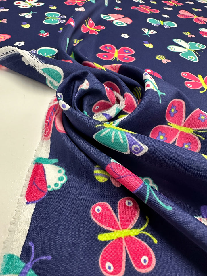 A piece of elegant dark blue fabric from Super Cheap Fabrics, named Printed Delsutered Satin - Butterfly - 155cm, is adorned with colorful butterfly patterns in pink, green, yellow, and red. The fabric is partly rolled at one end, revealing a neat edge.