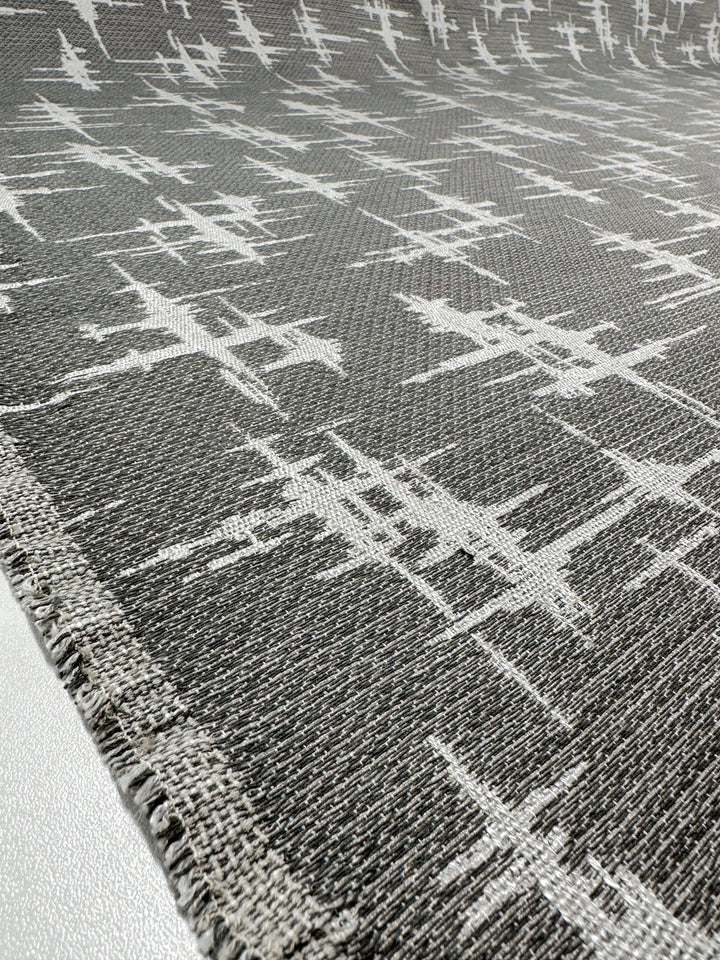 Close-up of the "Abstract Woven Upholstery - Morissey - 147cm" by Super Cheap Fabrics, featuring a gray textured upholstery fabric with an abstract white pattern of intersecting lines. Ideal for modern furniture, it showcases a contemporary weave and slightly frayed edge in the foreground.