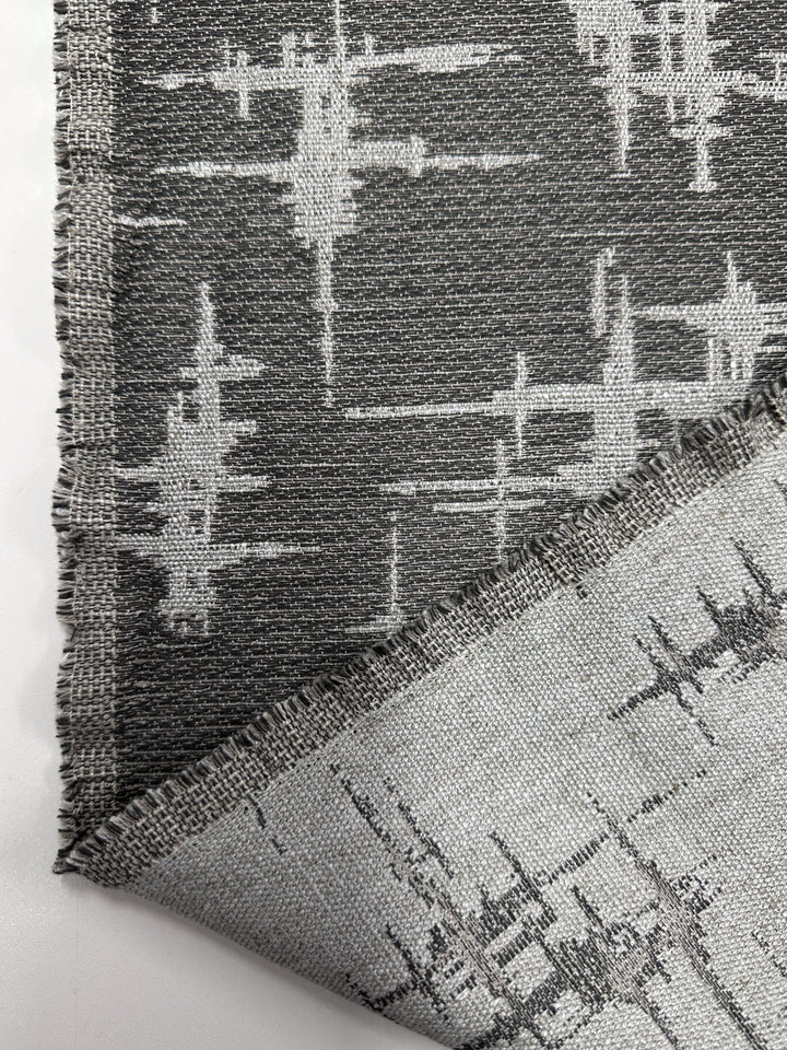 The Abstract Woven Upholstery - Morissey - 147cm by Super Cheap Fabrics features a gray and white abstract pattern, with one corner folded to reveal the mirrored reverse side. Its slightly frayed edges add a modern touch, ideal for contemporary furniture.