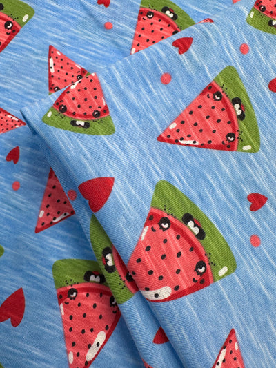 The Super Cheap Fabrics Printed Jersey - Sandia, 145cm wide, features a playful blue design with cartoon watermelon slices sporting smiling faces and small black seeds. Scattered red hearts add a cheerful charm, making it ideal for any project requiring delightful multi-color appeal.