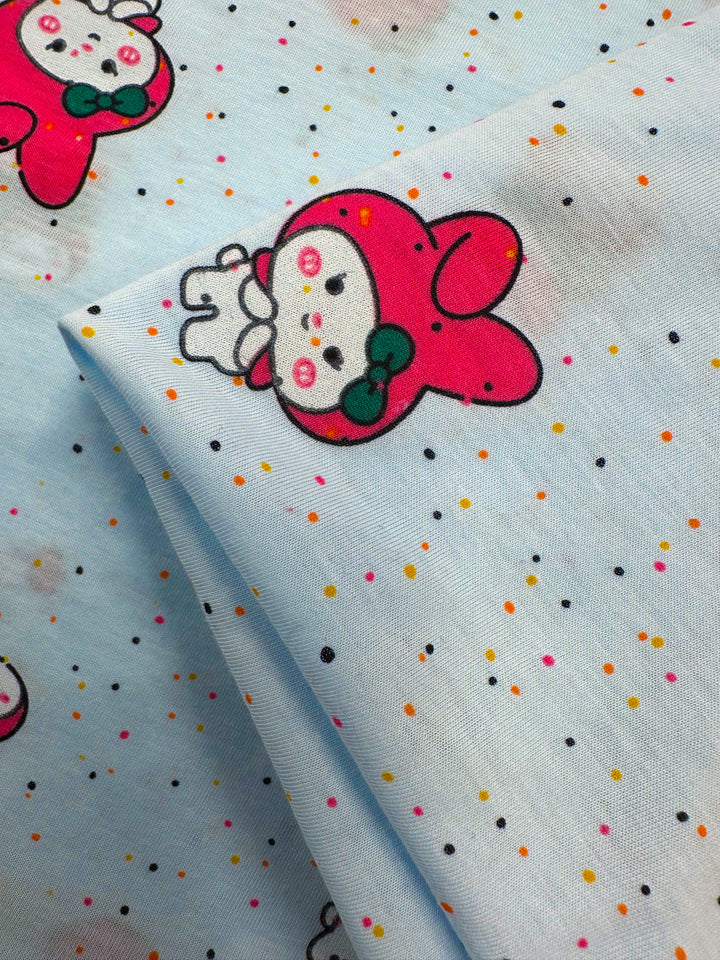 The Super Cheap Fabrics' Printed Jersey - Melody, 140cm, features adorable white bunnies with red hoods, green bows, and pink cheeks on a light blue backdrop adorned with small multicolored dots. This medium-weight fabric offers a playful touch with its two-way stretch design.