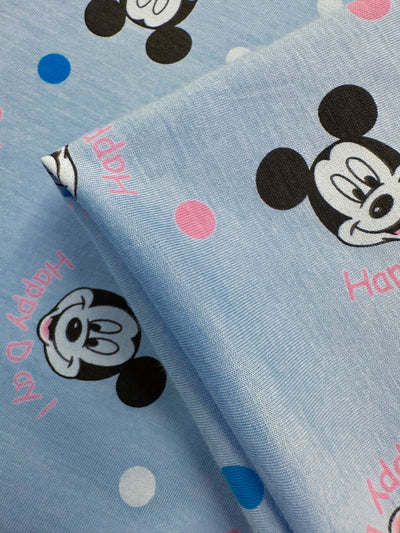 Close-up of Super Cheap Fabrics' "Printed Jersey - Cheers Ears - 150cm" featuring a cartoon mouse face pattern with multicolored polka dots and the phrase "Happy Day" in pink on light blue medium-weight fabric.