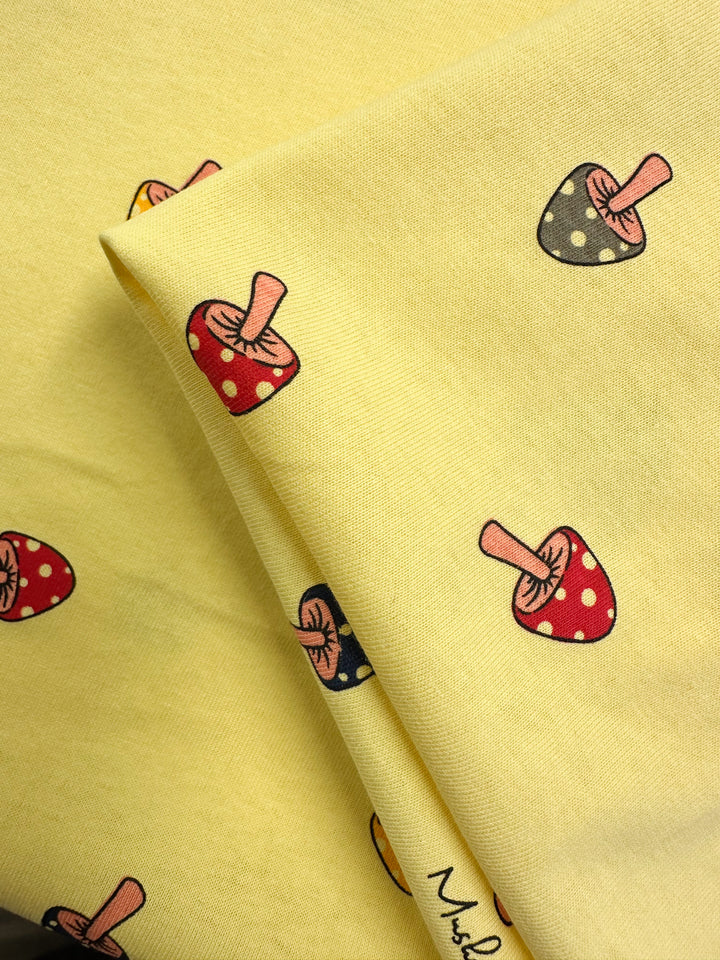 Super Cheap Fabrics' Printed Jersey - Shroom - 160cm features a yellow medium weight base with red and black mushrooms, white spots, and pink stems in a repeating pattern. The fabric’s folds highlight its vibrant, multi-color design.
