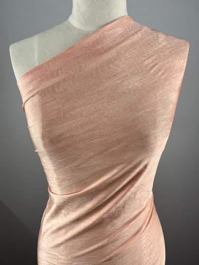 A mannequin displays Super Cheap Fabrics' Shimmer Lycra - Salmon - 150cm, a light pink, shimmery, one-shoulder fabric with gentle folds and a smooth, elegant texture against a neutral gray background.