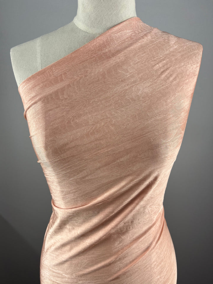 A mannequin displays Super Cheap Fabrics' Shimmer Lycra - Salmon - 150cm, a light pink, shimmery, one-shoulder fabric with gentle folds and a smooth, elegant texture against a neutral gray background.