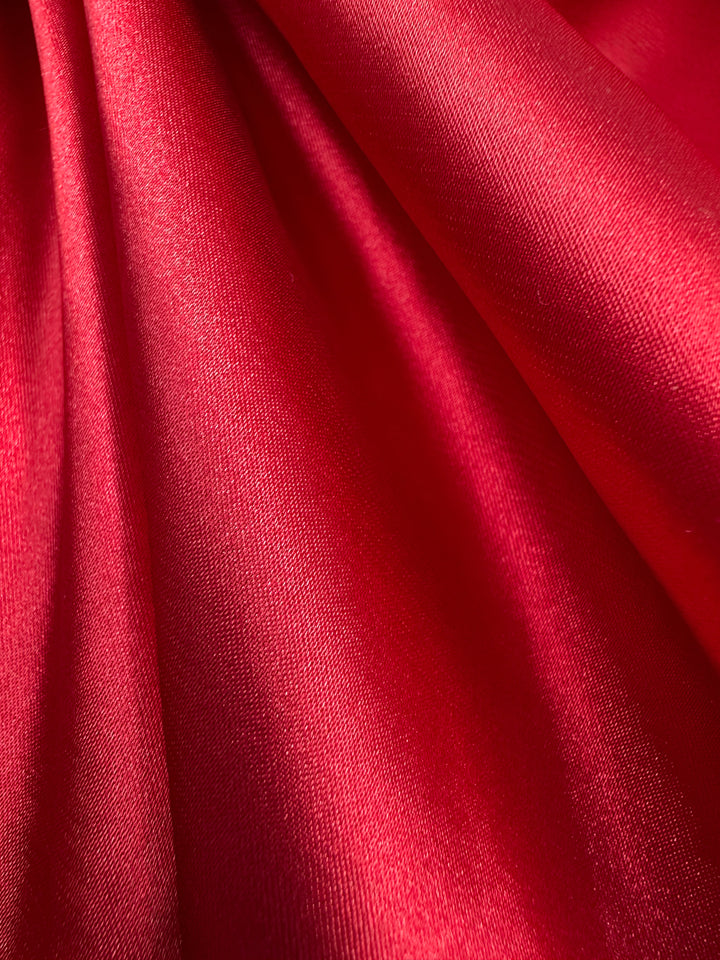 A close-up of the Satin Deluxe - Scarlet Smile - 150cm from Super Cheap Fabrics reveals its vibrant red satin silk with smooth, flowing folds and a lustrous sheen, highlighting the luxurious texture ideal for statement upholstery.