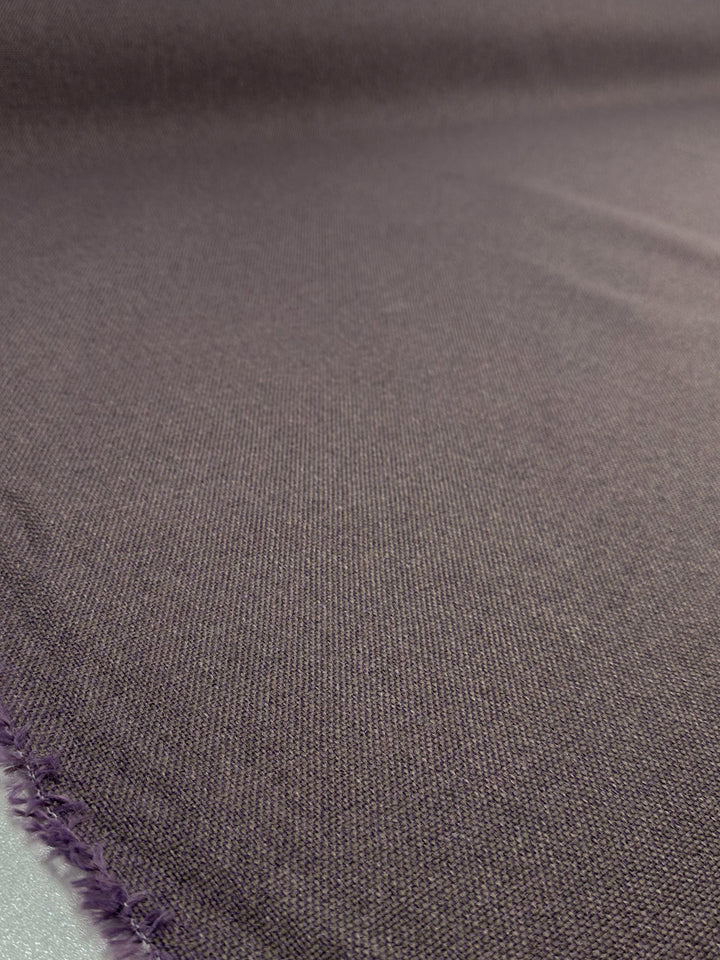 A close-up of the Two-Tone Weave Upholstery - Camden Dusk by Super Cheap Fabrics showcases a smooth surface, visible weaves, and a slightly frayed edge in the lower left corner. The lighting accentuates its dusty purple hue and texture variations.