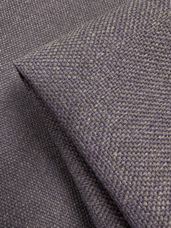 A close-up of dark gray, textured upholstery fabric reveals a two-tone weave in dusty purple. A folded section showcases the thickness and texture of Super Cheap Fabrics' Camden Dusk - 145cm.