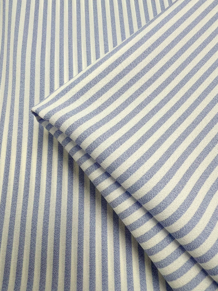 A close-up of Super Cheap Fabrics' Striped Crepe - Pale Iris (150cm), featuring blue and white vertical stripes with a folded section placed diagonally over the flat fabric background.