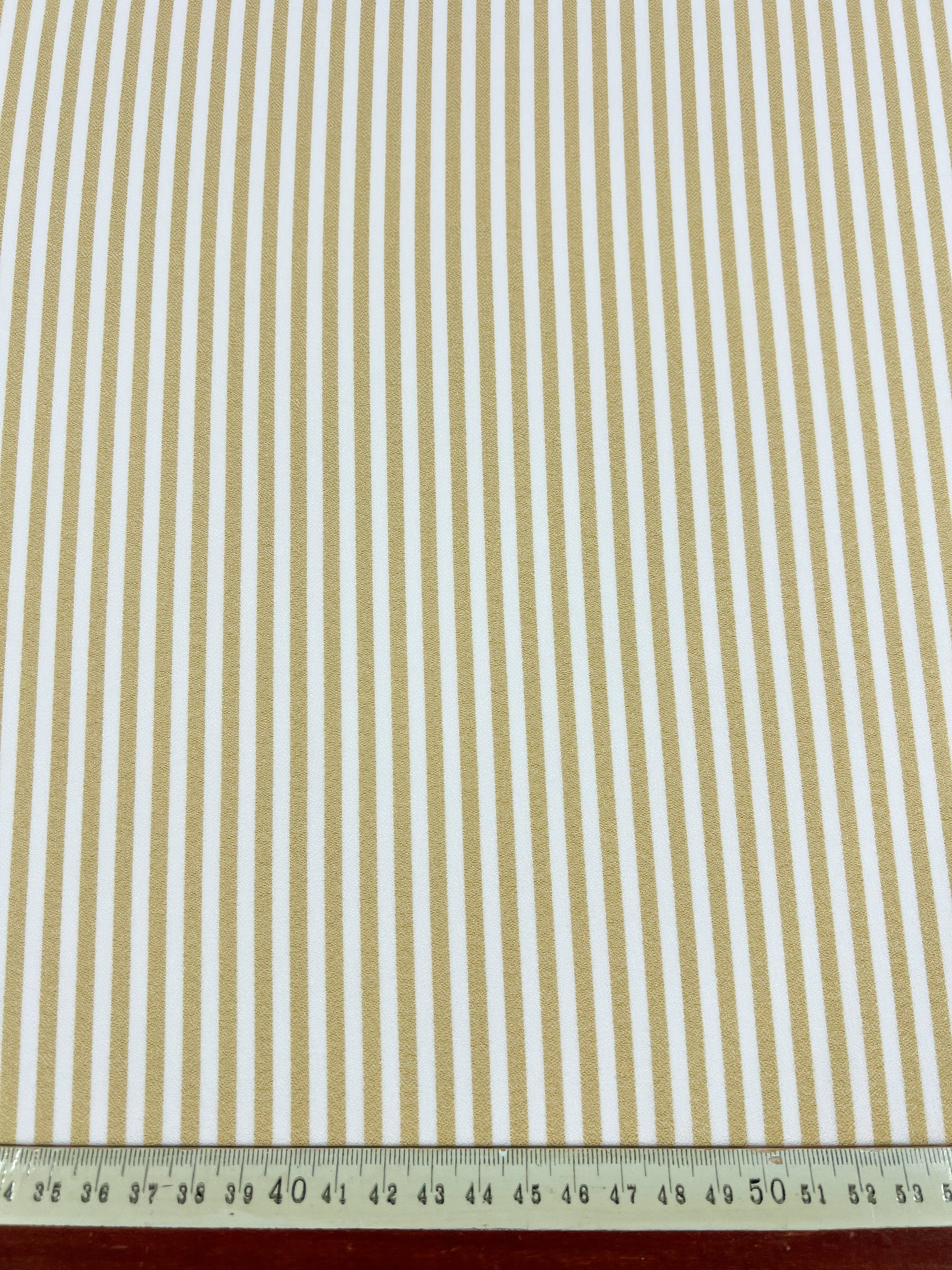The Super Cheap Fabrics Striped Crepe - Raffia - 150cm, a lightweight polyester fabric with vertical beige and white stripes, is displayed with a metal ruler at the bottom to show stripe width and overall scale.