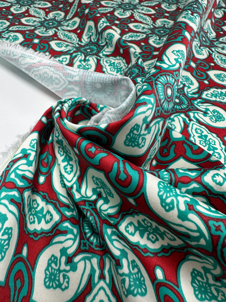 A close-up of Super Cheap Fabrics’ Cotton Sateen - Mural - 150cm shows its lightweight fabric with vibrant teal, red, and white abstract floral and geometric designs—ideal for home décor—partially rolled and draped flat.