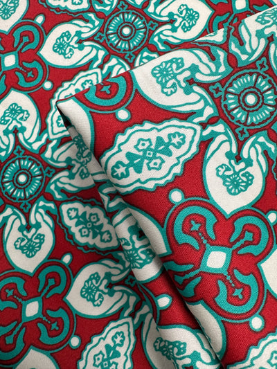 Super Cheap Fabrics' Cotton Sateen - Mural - 150cm is a red lightweight fabric with teal and white geometric and floral designs, perfect for home décor. The fabric is pictured folded in the foreground.