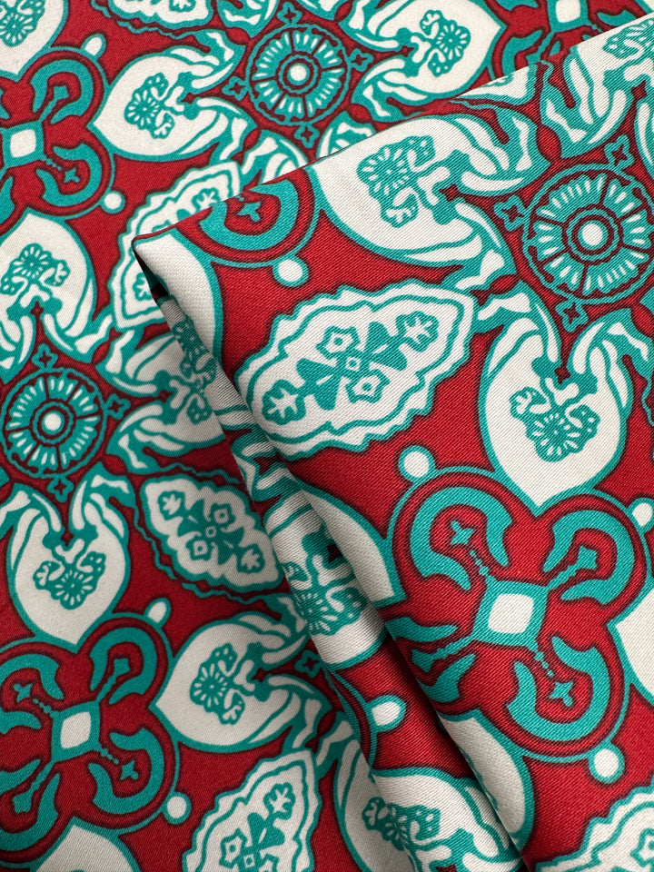 Super Cheap Fabrics' Cotton Sateen - Mural - 150cm is a red lightweight fabric with teal and white geometric and floral designs, perfect for home décor. The fabric is pictured folded in the foreground.