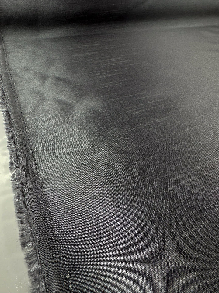 A close-up of Super Cheap Fabrics' Shantung Satin - Charcoal (150cm) shows its smooth, shiny surface, subtle texture, neat edge stitching, and a slightly frayed selvage on the left side.