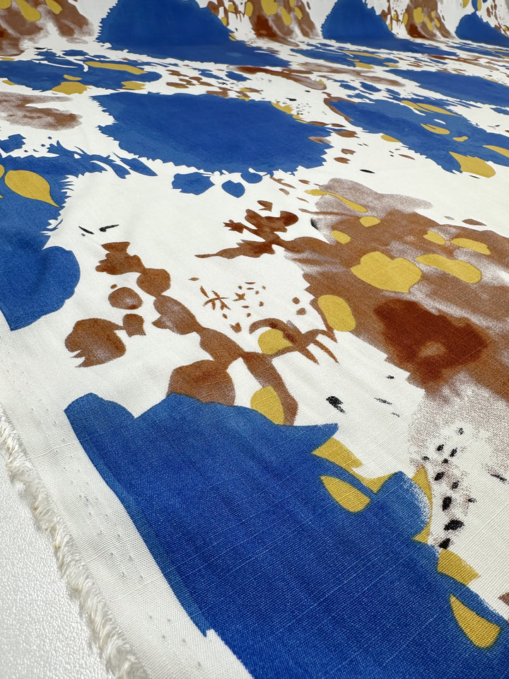 A close-up of Bamboo Rayon - Splash - 150cm by Super Cheap Fabrics shows lightweight fabric with an abstract blue, brown, yellow, and white painterly splatter design and frayed edges—ideal for sewing projects.