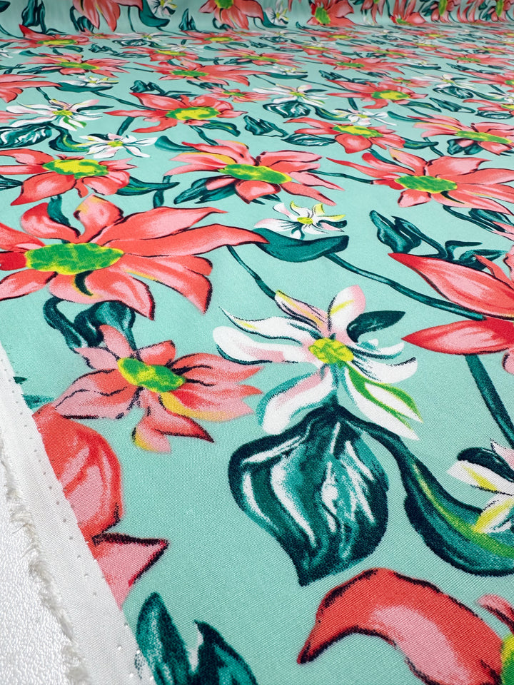 Close-up of Super Cheap Fabrics' Designer Rayon - Tropical Blossom (145cm), showcasing vibrant red, pink, and white floral prints with green leaves on aqua fabric, featuring a bold repeating pattern.