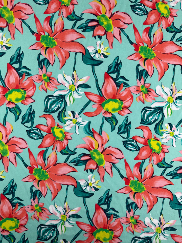 Designer Rayon - Tropical Blossom by Super Cheap Fabrics features large red and white flowers with green leaves and yellow centers on a teal background. The 145cm versatile rayon offers bold, vibrant prints with densely packed floral designs.