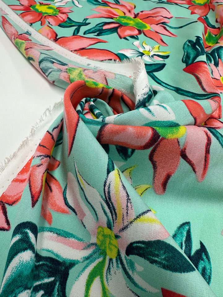 A close-up of Super Cheap Fabrics’ Designer Rayon - Tropical Blossom (145cm), showing colorful floral patterns with large pink, white, and yellow flowers plus green leaves on light turquoise, partially folded to reveal texture and selvage.