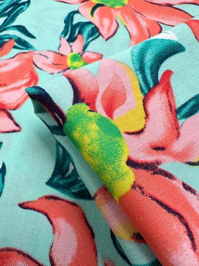 A close-up of Super Cheap Fabrics' Designer Rayon - Tropical Blossom (145cm), featuring vibrant coral, pink, green, and yellow florals on light blue. A corner is folded to show the premium fabric's underside.