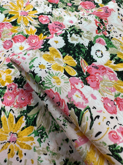 A close-up of Designer Rayon - Floral Garden by Super Cheap Fabrics shows vibrant yellow, pink, and white flowers with green leaves on a light background. Its lively print and soft drape make it a versatile 145cm-wide fabric for many projects.