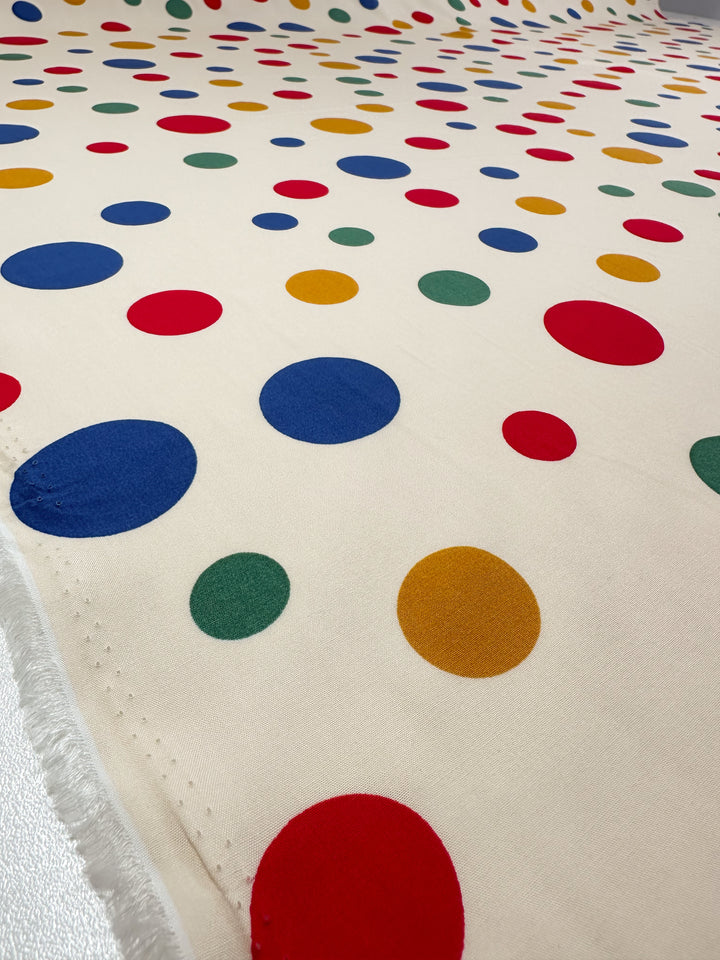 Super Cheap Fabrics’ Designer Rayon - Pop of Colour - 145cm is a cream fabric with vivid red, blue, green, and yellow polka dots. Slightly wrinkled with frayed edges visible on the left side.