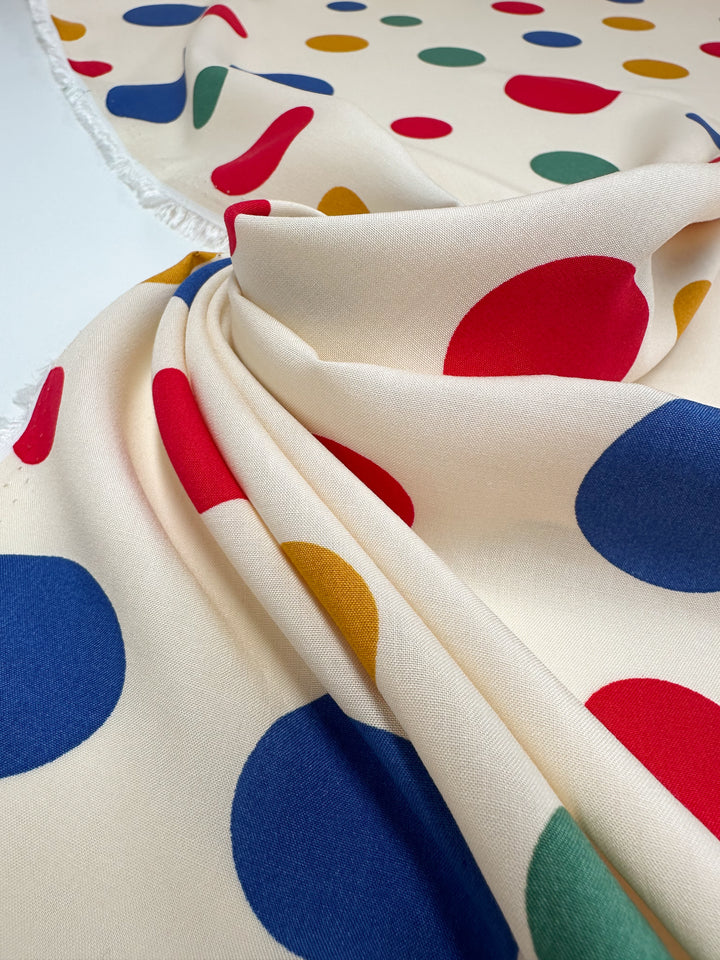 A close-up of Super Cheap Fabrics' Designer Rayon - Pop of Colour (145cm), featuring cream fabric with large, vibrant red, blue, yellow, and green polka dots. The versatile material drapes softly with a textured surface and unfinished edges.