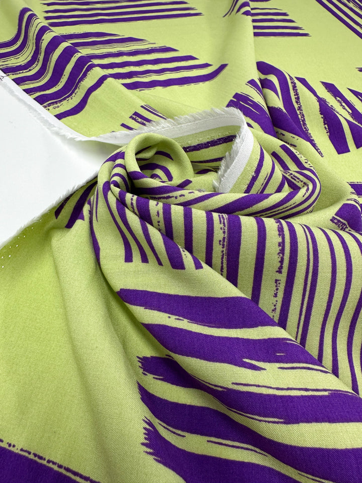 A close-up of Designer Rayon - Geometric Stripes by Super Cheap Fabrics in light green with bold purple brushstroke stripes, partially rolled and gathered—perfect for vibrant, eye-catching creations. Width: 145cm.