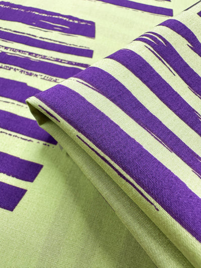 A close-up of Super Cheap Fabrics’ Designer Rayon – Geometric Stripes (145cm) in beige, showcasing bold, irregular purple stripes and brushstroke patterns with a folded section highlighting its textured weave.