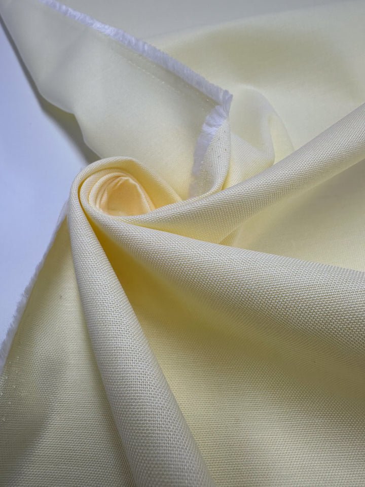 A close-up of Super Cheap Fabrics' Oxford Cotton - Lemon (146cm) shows its pale yellow hue and subtle texture, loosely gathered into a swirl to display both the smooth surface and unfinished white edge.