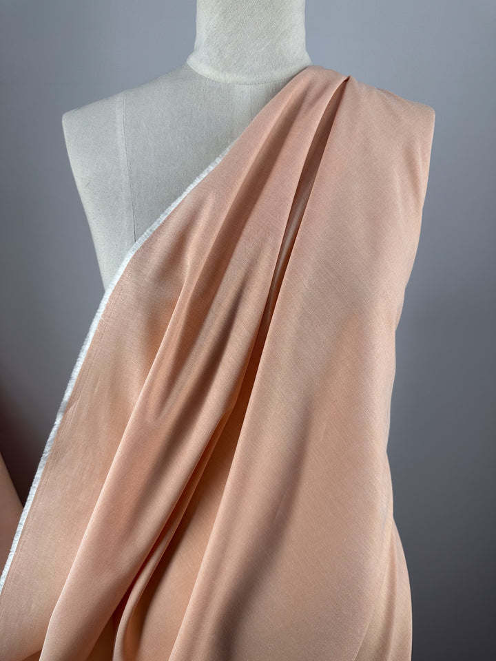 A mannequin showcases Super Cheap Fabrics’ Oxford Cotton - Sunburst - 146cm in light peach with a subtle sheen, draped diagonally to leave one shoulder exposed, set against a plain, neutral background.