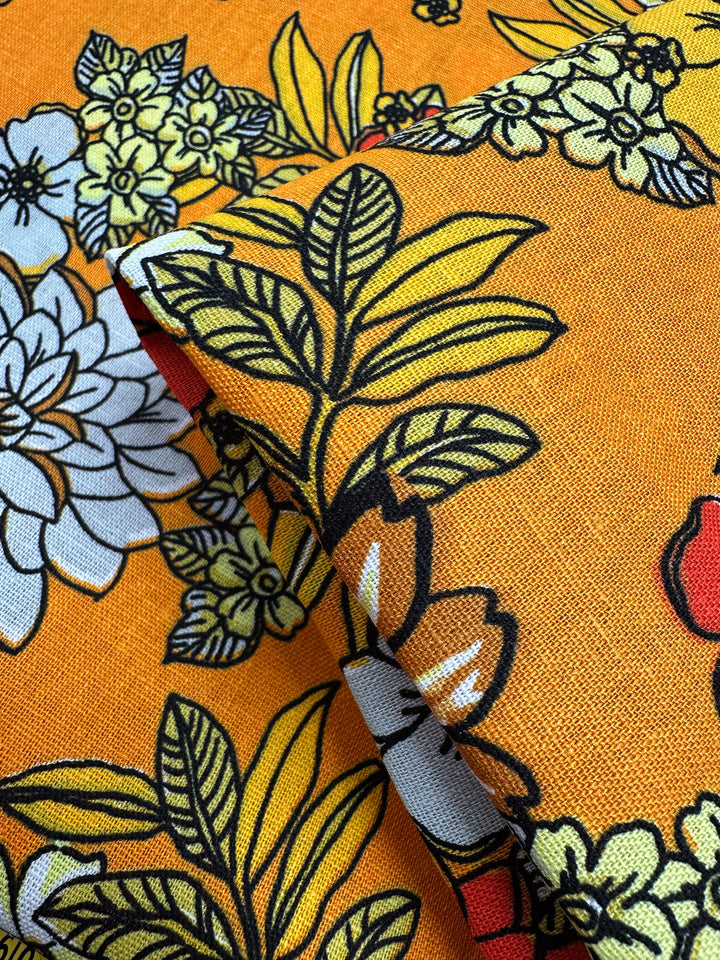 The Printed Linen Blend - Golden Garden by Super Cheap Fabrics is a bright orange medium-weight linen blend with bold yellow, white, and black floral patterns, featuring large leaves and flowers. Fabric is shown folded to highlight texture and design.