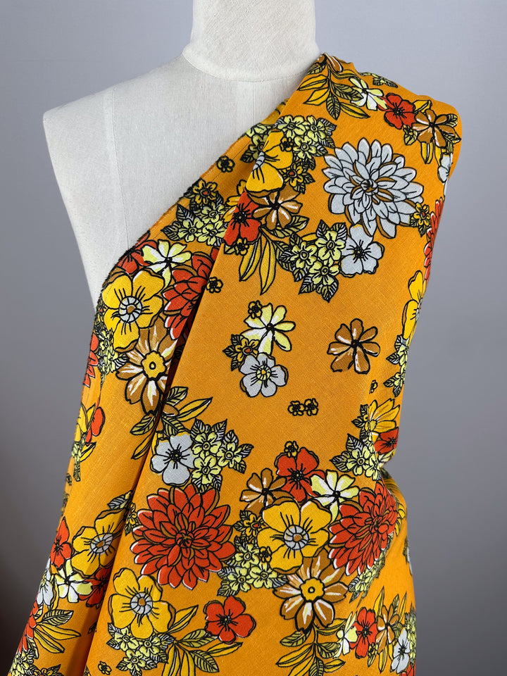 A white mannequin showcases Super Cheap Fabrics' Printed Linen Blend - Golden Garden (128cm), a medium-weight linen with a bold floral print in red, orange, yellow, and white flowers with green leaves on bright yellow, against a gray background.