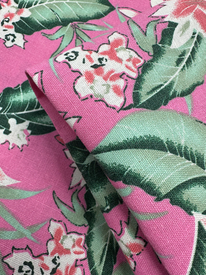 Close-up of Super Cheap Fabrics' Printed Linen Blend - Jungle Bloom (140cm), showing pink fabric with a tropical floral pattern—large green leaves and white-pink flowers. The medium weight linen cotton blend is partially folded for added texture.