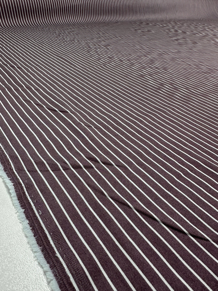 A close-up of the Super Cheap Fabrics Chambray - Grape Shake (150cm), showing maroon fabric with white vertical stripes and a frayed left edge, made from lightweight cotton nylon blend and laid flat.