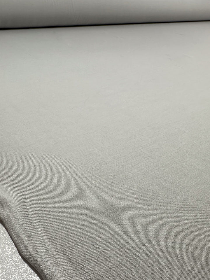 A close-up of Super Cheap Fabrics' Cotton Lycra - Moonstruck (168cm), light gray with a smooth finish and subtle stretch texture, rolled out flat with the fabric roll visible at the top.