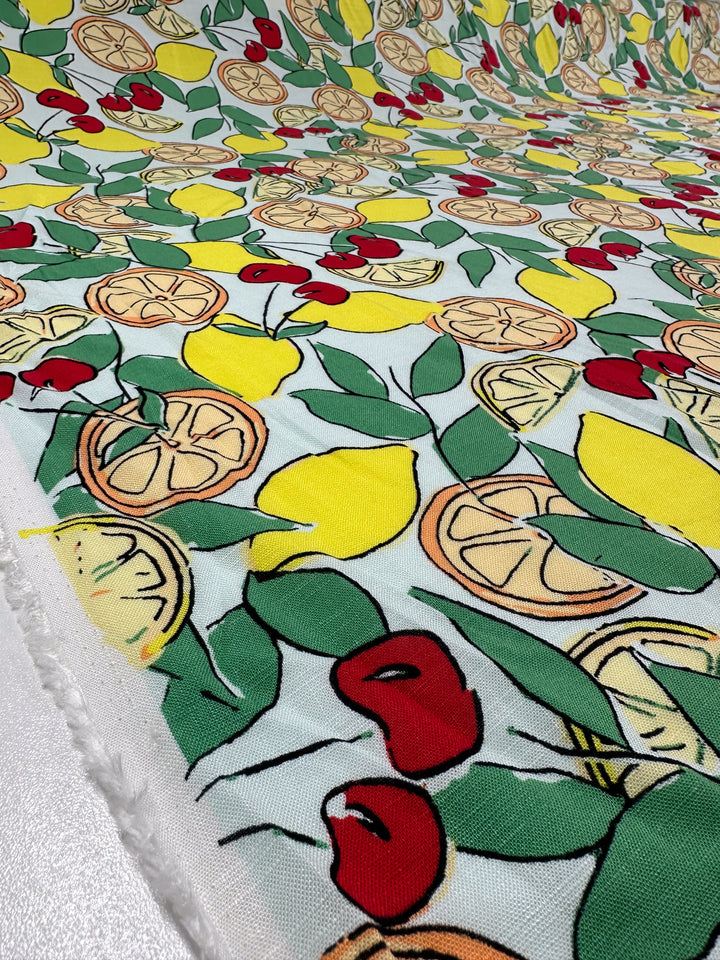 Close-up of Super Cheap Fabrics’ Printed Linen Blend - Citrus Crush (134cm) showing lemons, lemon slices, cherries, and green leaves on a light background—ideal for upholstery or vibrant summer jackets.