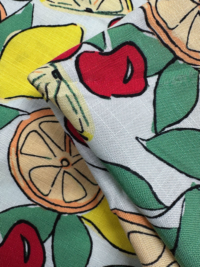 Close-up of Super Cheap Fabrics' Printed Linen Blend - Citrus Crush (134cm), featuring lemons, oranges, cherries, and green leaves on a light background—ideal for summer jackets or upholstery. Fabric is slightly folded in one corner.