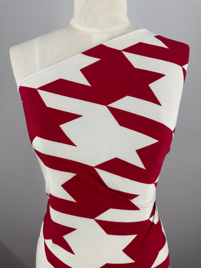 A mannequin showcases Super Cheap Fabrics’ Printed Lycra - Bold Bite (154cm), featuring striking red zigzag and arrow-inspired geometric prints on white in a one-shoulder style, set against a plain gray background.