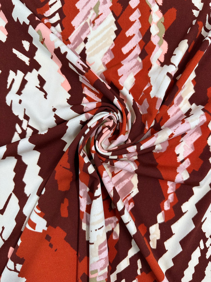 A close-up of Super Cheap Fabrics' Printed Lycra - Crimson Crush (150cm), medium weight fabric with a swirling twist and a bold zigzag pattern in red, burgundy, white, pink, and light green.