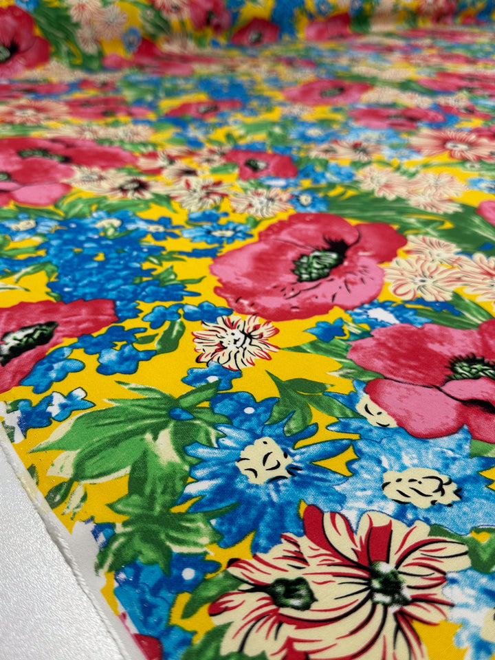 A close-up of Super Cheap Fabrics’ Designer Rayon - Sunset Bloom (145cm), featuring bold red, blue, and white floral prints with green leaves densely arranged on a vibrant yellow background for an eye-catching look.