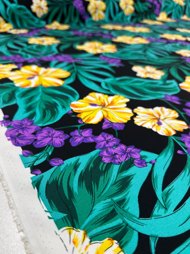 A close-up of Super Cheap Fabrics' Designer Rayon - Neon Canopy (145cm), displaying vibrant yellow and purple tropical flowers with green leaves on a black background. The fabric edge highlights its lightweight rayon quality.