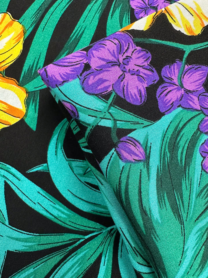 Close-up of Designer Rayon - Neon Canopy (145cm) by Super Cheap Fabrics, showing lightweight rayon with a vibrant floral pattern in yellow, purple, and green on black, with fabric pieces artfully folded.